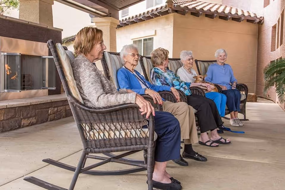 Five elderly women sitting in rocking chairs on a covered patio outside a building, with one woman holding a small dog. There is a stone fireplace with a fire burning on the left side and some greenery on the right side.