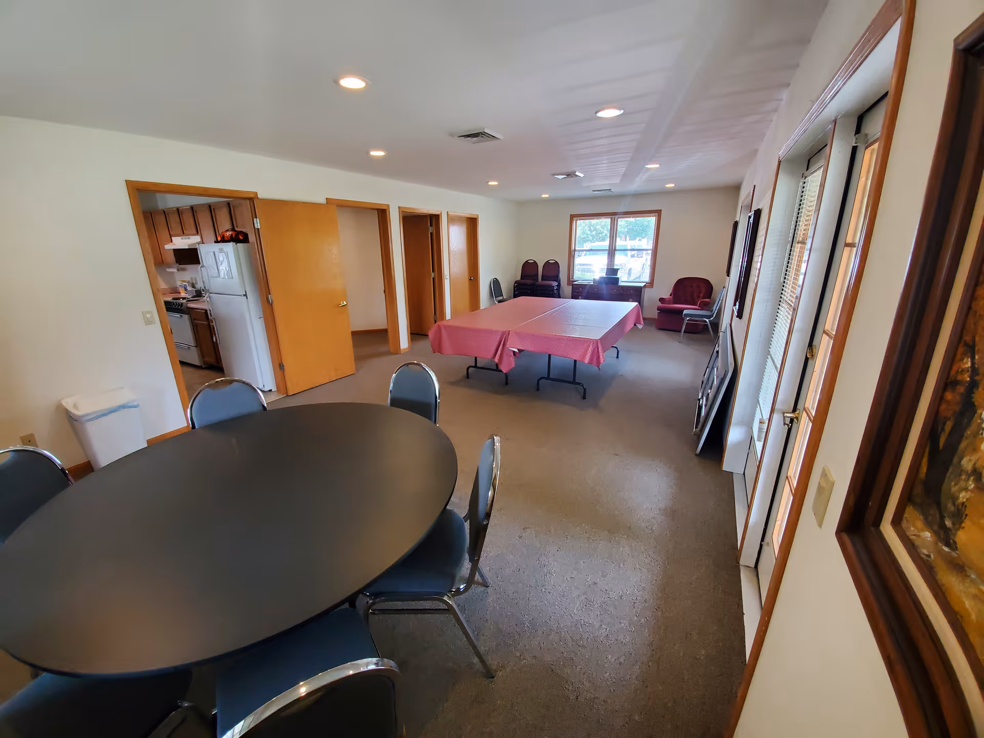 Interior room with a round black table surrounded by chairs in the foreground, a rectangular table covered with a pink tablecloth in the middle, and a kitchen visible through an open door on the left. There are several closed wooden doors along the back wall, a window with natural light, and some stacked chairs and an armchair near the window. The room has white walls, recessed ceiling lights, and carpeted flooring.