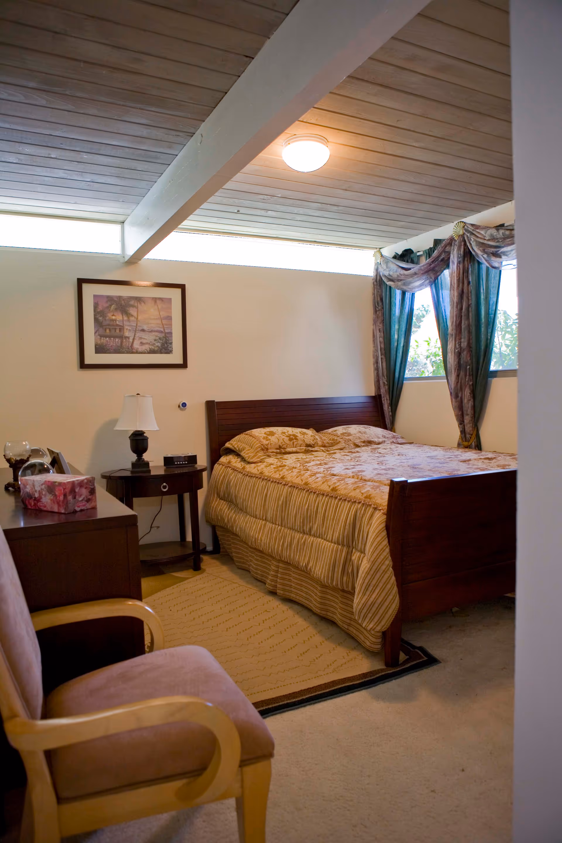 A cozy bedroom with a wooden bed, patterned bedding, nightstand and lamp, upholstered chair, and a curtained window.