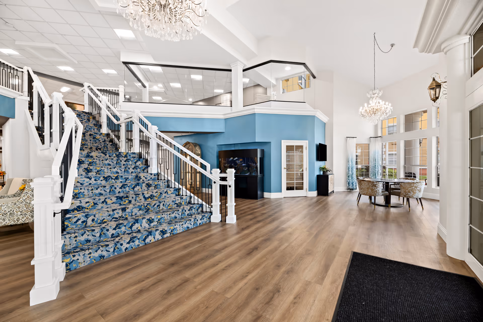 Bright and spacious interior of a senior living facility featuring a wide staircase with blue patterned carpet, white railings, and a large chandelier hanging from the ceiling. The room has wood flooring, a blue accent wall with a large fish tank, a round table with four upholstered chairs near tall windows, and decorative columns.