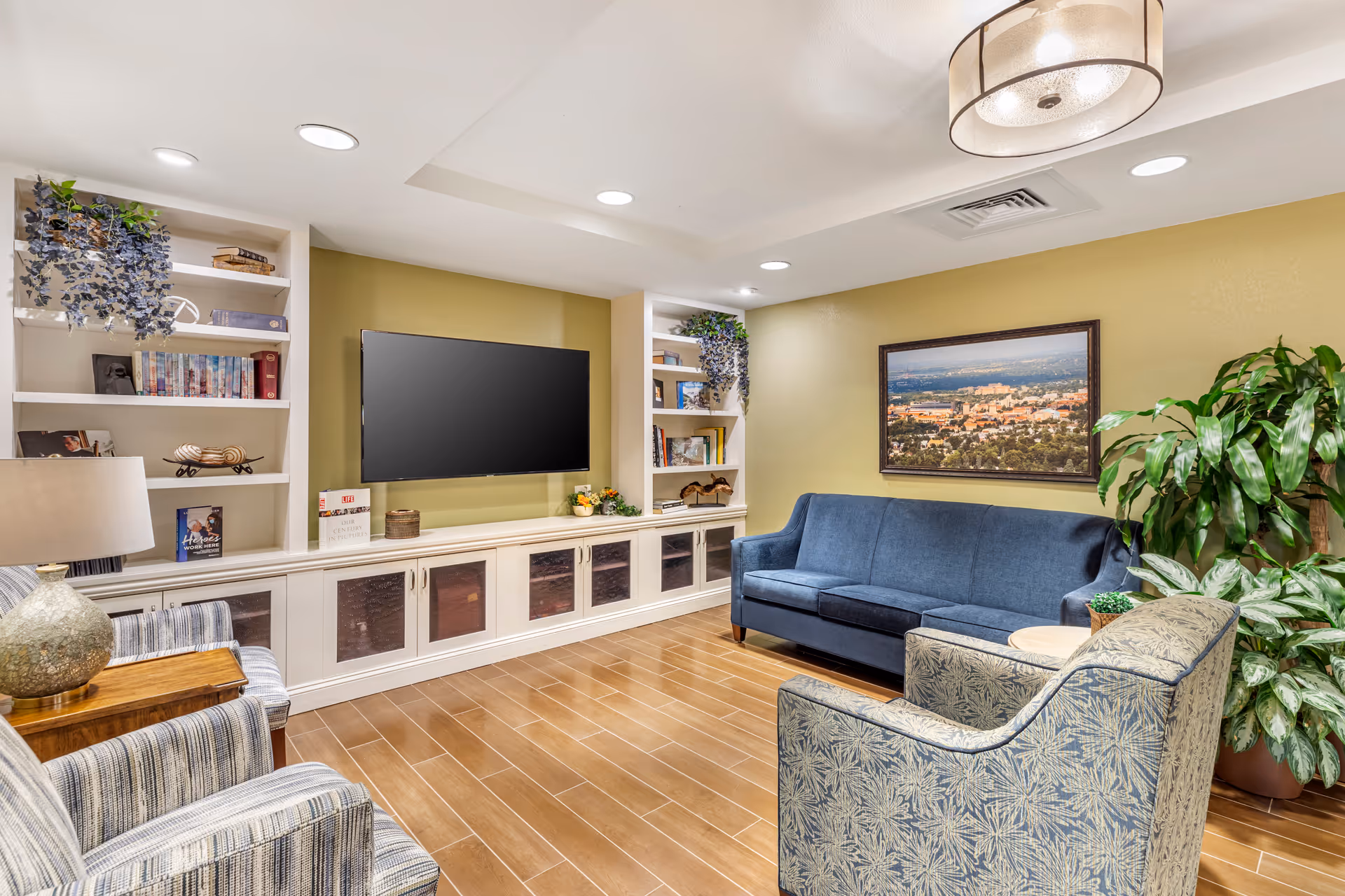 A cozy living room with a blue sofa, two patterned armchairs, a wooden side table with a lamp, built-in white shelves with books and decorative items, a flat-screen TV mounted on a green accent wall, a framed cityscape picture, and several potted plants. The room has wood-look tile flooring and recessed ceiling lights.