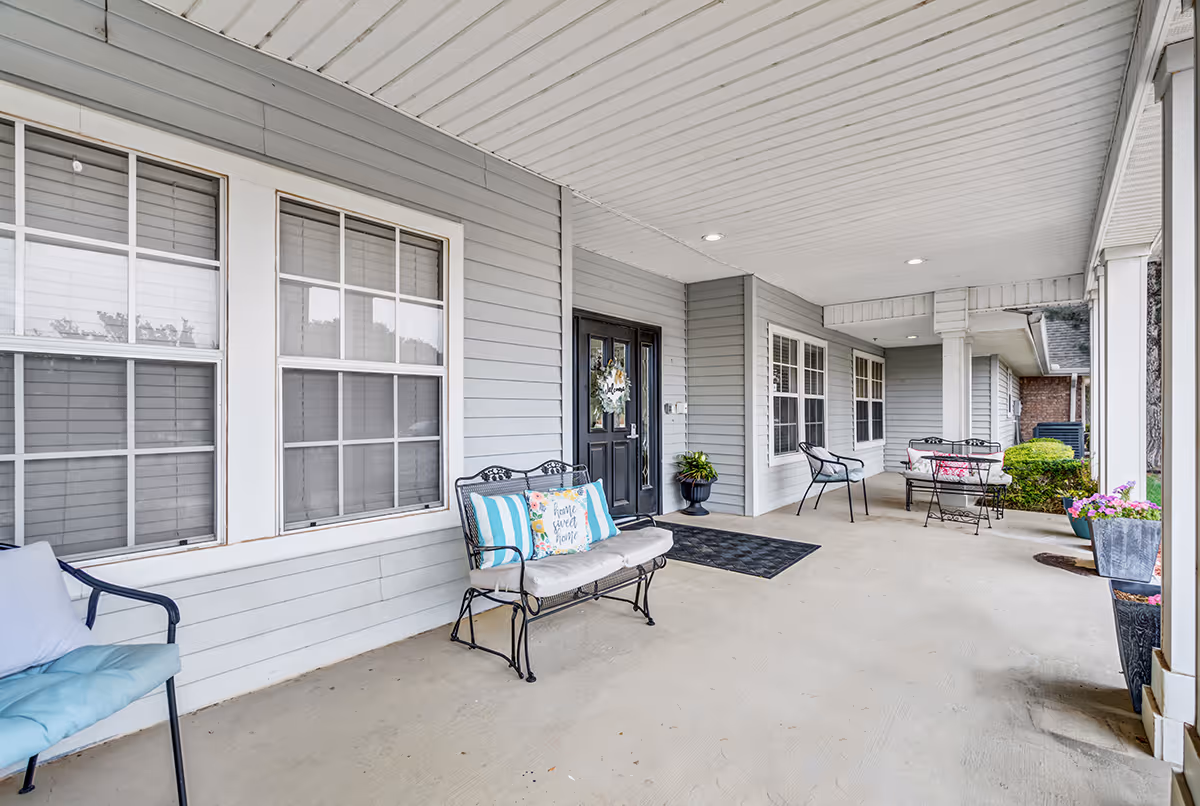 Covered porch area with gray siding walls and white ceiling. Several metal chairs and benches with cushions and pillows are arranged along the porch. A black door with a decorative wreath is visible, along with potted plants and flowers near the entrance.