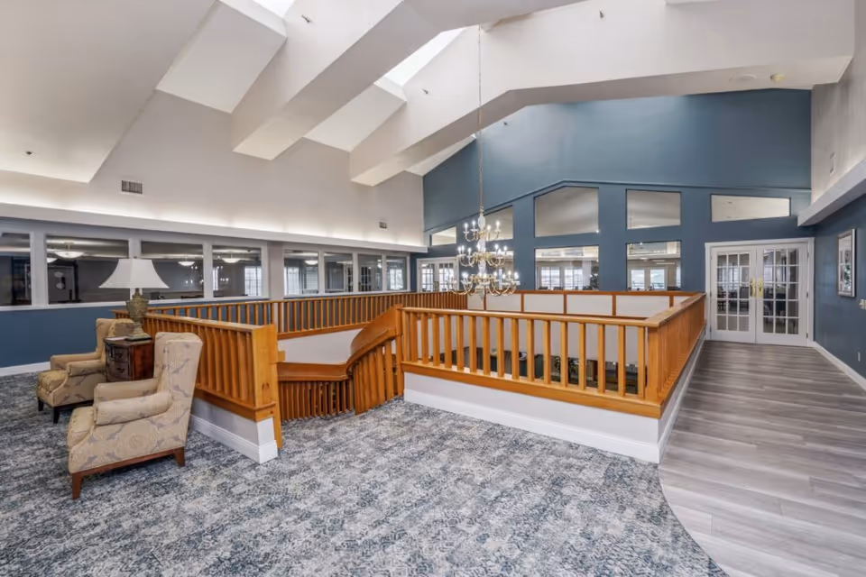 Interior view of a senior living facility common area with a wooden staircase railing surrounding an open stairwell. The space features patterned carpet flooring, two upholstered armchairs with a side table and lamp, blue accent walls, large windows, and a chandelier hanging from the ceiling with skylights above.