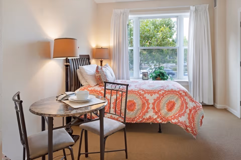 A cozy bedroom with a single bed covered in a colorful patterned bedspread in shades of orange and beige. There are two pillows on the bed and a bedside table with a lamp. Next to the bed is a small round table with two chairs, set with a cup and saucer on a napkin. A large window with white curtains lets in natural light and shows green trees outside.