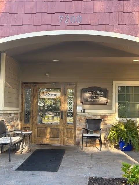 Entrance to a building with wooden double doors, a welcome sign on the door, and a plaque reading 'Nancy's Place 7200 SW Uplands Circle Aldersgate Village Life Plan Community'. There are two chairs and a small table on the left side, a chair and a large potted plant on the right side, and the number 7200 above the entrance.