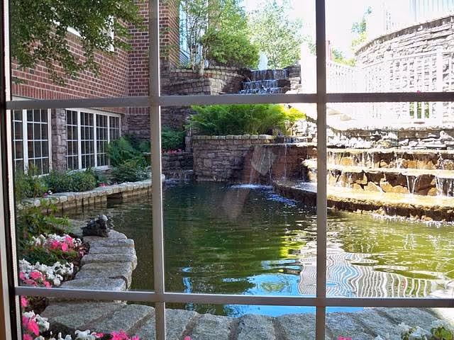 View through a window showing a serene outdoor water feature with a pond, stone walls, cascading waterfalls, and surrounding greenery and flowers at Bethel Health Care Center.
