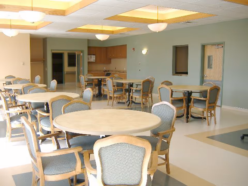 A bright dining room with multiple round tables surrounded by wooden chairs with cushioned seats and backs. The room has a light-colored floor with patterned tiles, green walls, and a ceiling with recessed lighting and hanging light fixtures. There is a small kitchenette area with cabinets and a sink in the background.