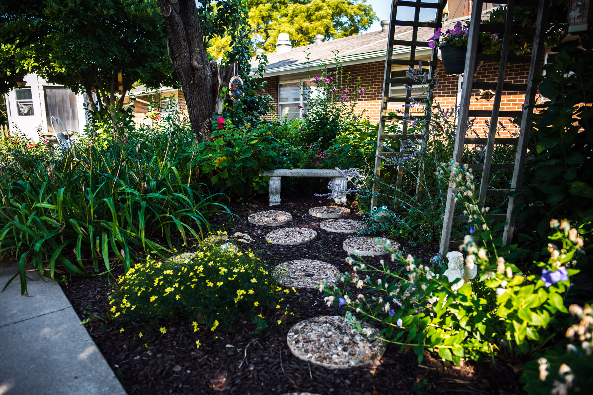 A lush garden area with a stone pathway made of circular stepping stones leading to a small stone bench. The garden is filled with various green plants, flowers, and a tree providing shade. A brick building is visible in the background.