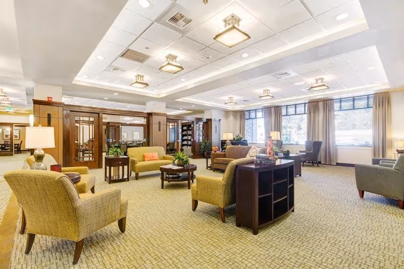 A spacious and well-lit living room area in a senior living facility with multiple armchairs and sofas arranged around coffee tables. The room features large windows with curtains, carpeted floors, and ceiling lights. Wooden partitions and shelves with plants and books are visible in the background.