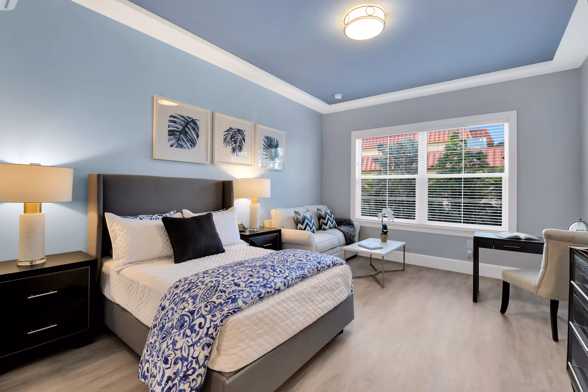 A bright and modern bedroom with a large window letting in natural light. The room features a queen-sized bed with white and blue bedding, two bedside tables with lamps, a small white couch with patterned pillows, a coffee table, and a desk with a chair. The walls are painted light blue with three framed leaf prints above the bed.