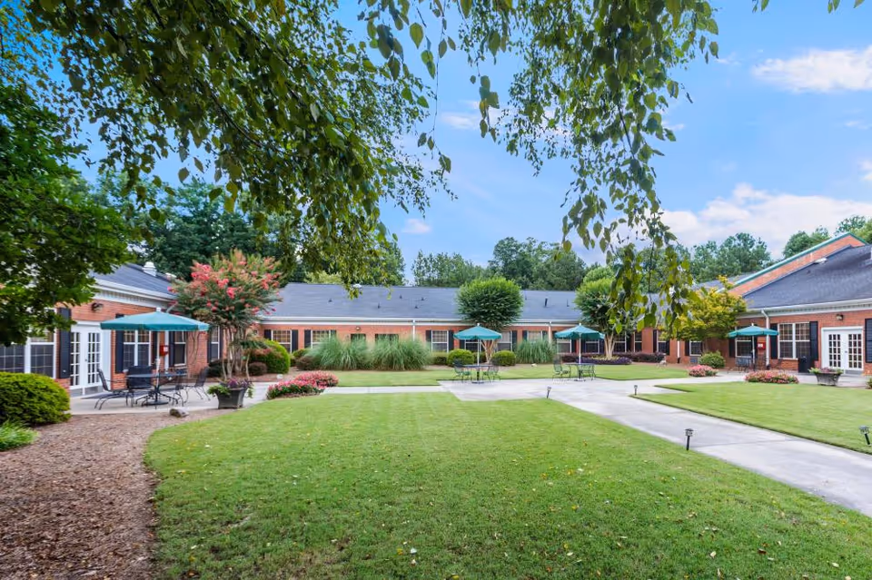 Outdoor courtyard area of Sandy Springs Center for Nursing and Healing with green grass, trees, shrubs, and several patio tables with umbrellas. The courtyard is surrounded by a single-story brick building with multiple windows and doors.