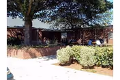 Exterior view of a single-story brick building with a shaded patio area. There are bushes and a large tree providing shade near the entrance. A few people are sitting on benches under the covered patio.