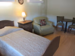 A bedroom with a single bed covered in a light-colored quilt, a wooden nightstand with a lamp and a clock on the wall above it, a cushioned armchair with a decorative pillow, and a small round table with two wooden chairs in the background.