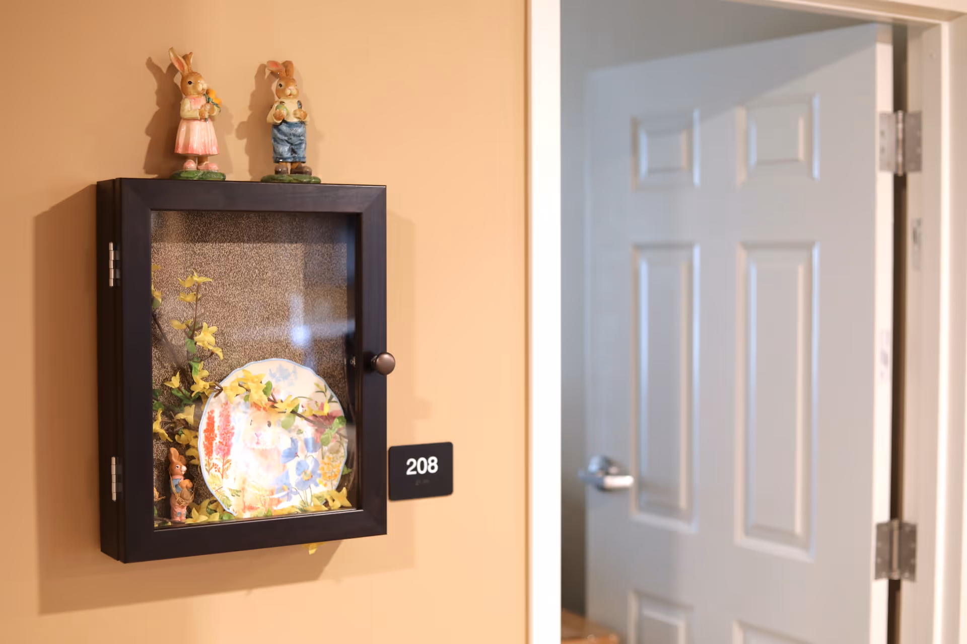A beige wall with a small black display cabinet mounted on it, containing a decorative plate and artificial yellow flowers. Two ceramic bunny figurines are placed on top of the cabinet. To the right, a partially open white door with a silver handle is visible. A black room number plaque with the number 208 is mounted on the wall next to the cabinet.