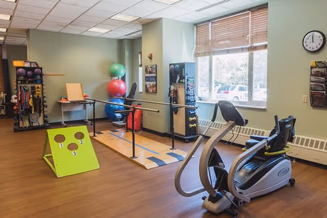 A well-lit exercise room with wooden flooring featuring various fitness equipment including an exercise bike, balance balls stacked in a corner, a green cornhole board, resistance bands, and parallel bars for walking support. Large windows with blinds allow natural light to enter the room.