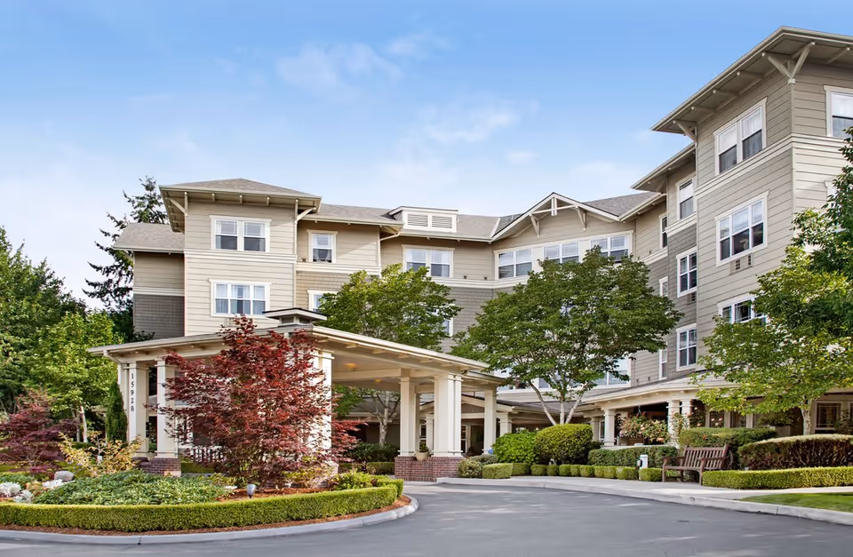 Exterior view of a multi-story senior living facility with beige siding, multiple windows, a covered entrance with white columns, landscaped greenery, and a clear blue sky.