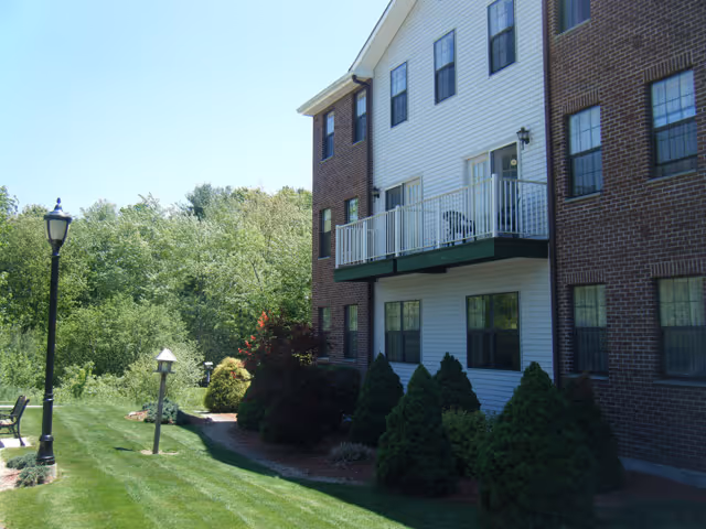 Side view of a multi-story brick-and-siding senior living building with a balcony, manicured lawn, lamp posts, and trees.