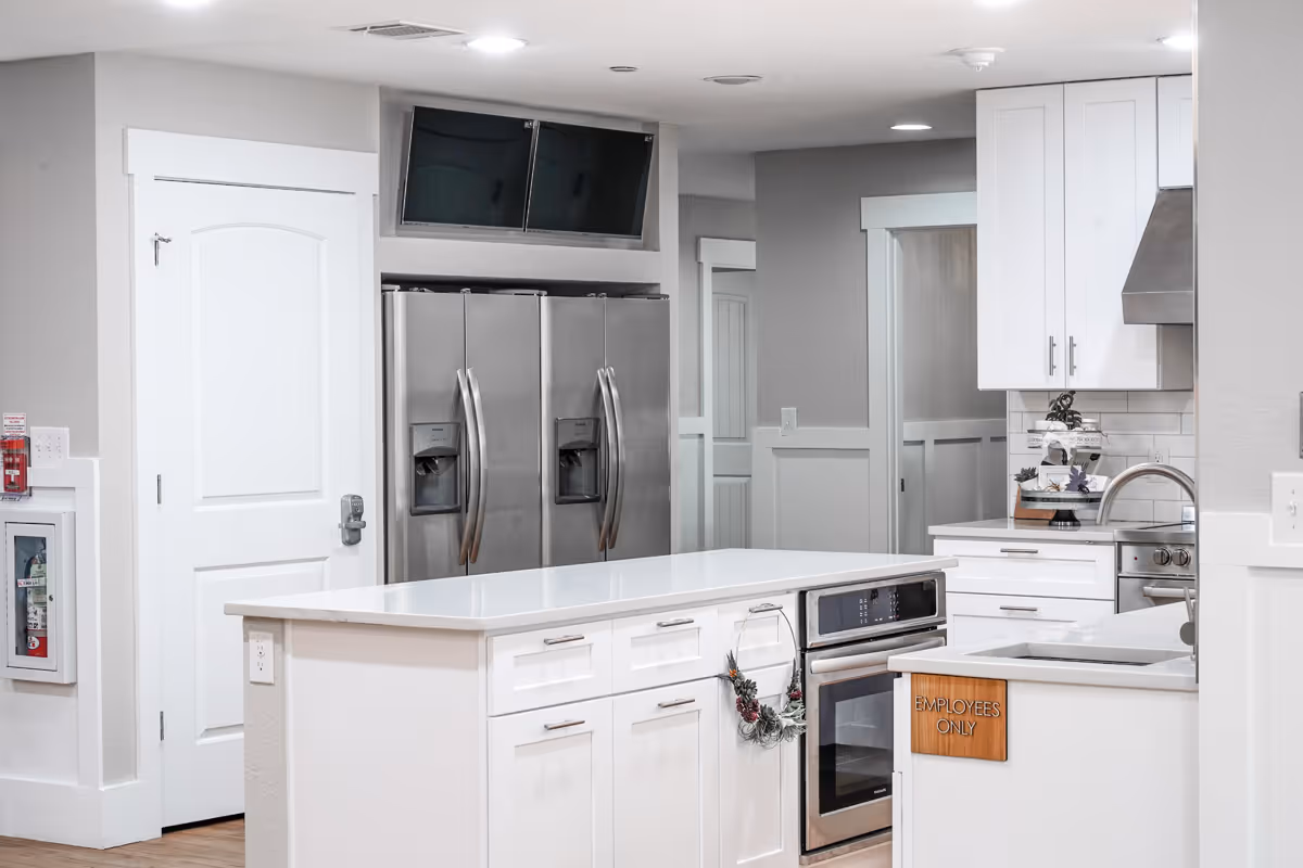 A modern kitchen area with white cabinetry, a large white island countertop, stainless steel double-door refrigerators, an oven, and a sink. There is a wooden sign on the counter that reads 'EMPLOYEES ONLY'. The walls are light gray with white trim, and there are two black screens mounted above the refrigerators.