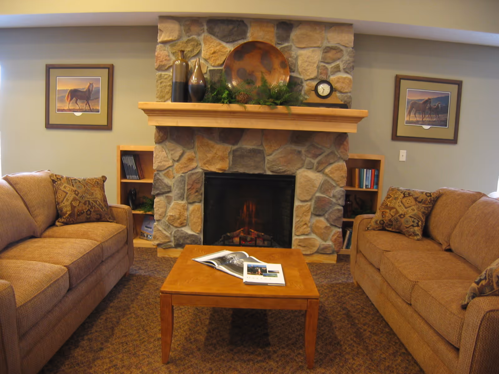 A cozy living room with two brown sofas facing each other across a wooden coffee table with an open magazine on it. In the background, there is a stone fireplace with a wooden mantel decorated with vases, a round decorative plate, greenery, and a small clock. On either side of the fireplace are built-in wooden shelves with books and decorative items. Two framed pictures of horses hang on the walls above the sofas.