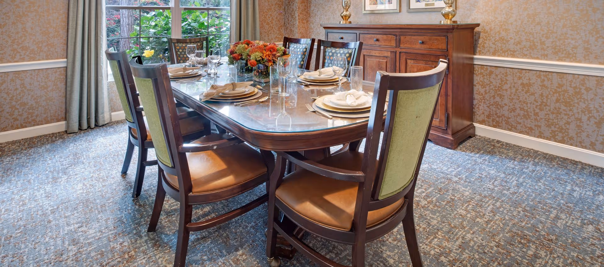 A dining room with a wooden table set for six, upholstered chairs, a floral centerpiece, sideboard, and a large window overlooking greenery.