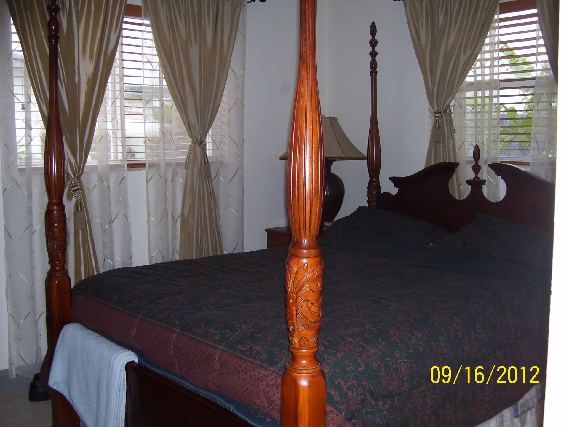 A bedroom with a dark wooden four-poster bed covered with a dark patterned bedspread. The room has two windows with beige curtains and sheer white drapes. A bedside table with a lamp is visible next to the bed.