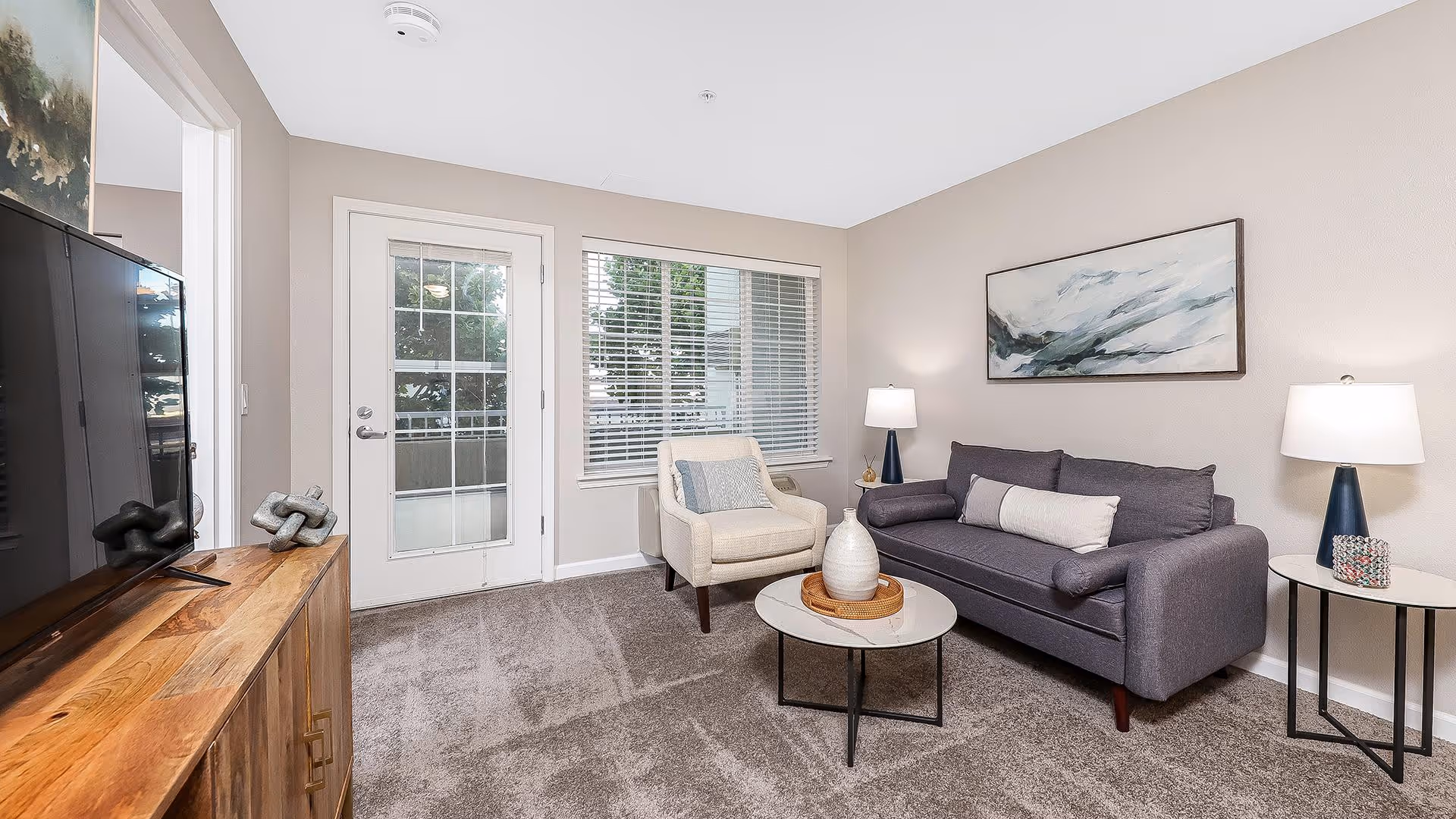 A cozy living room with a gray sofa, a white armchair, a round coffee table with decorative items, two side tables with lamps, a large flat-screen TV on a wooden cabinet, and a window and glass door leading outside.