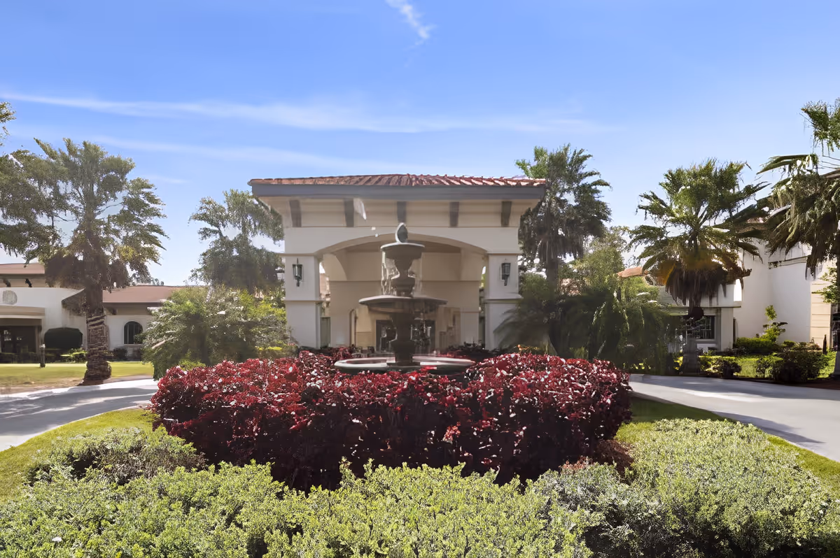 Front exterior view of Discovery Commons South Biscayne featuring a circular driveway with a central fountain surrounded by red and green shrubs, palm trees, and a building with a tiled roof under a clear blue sky.