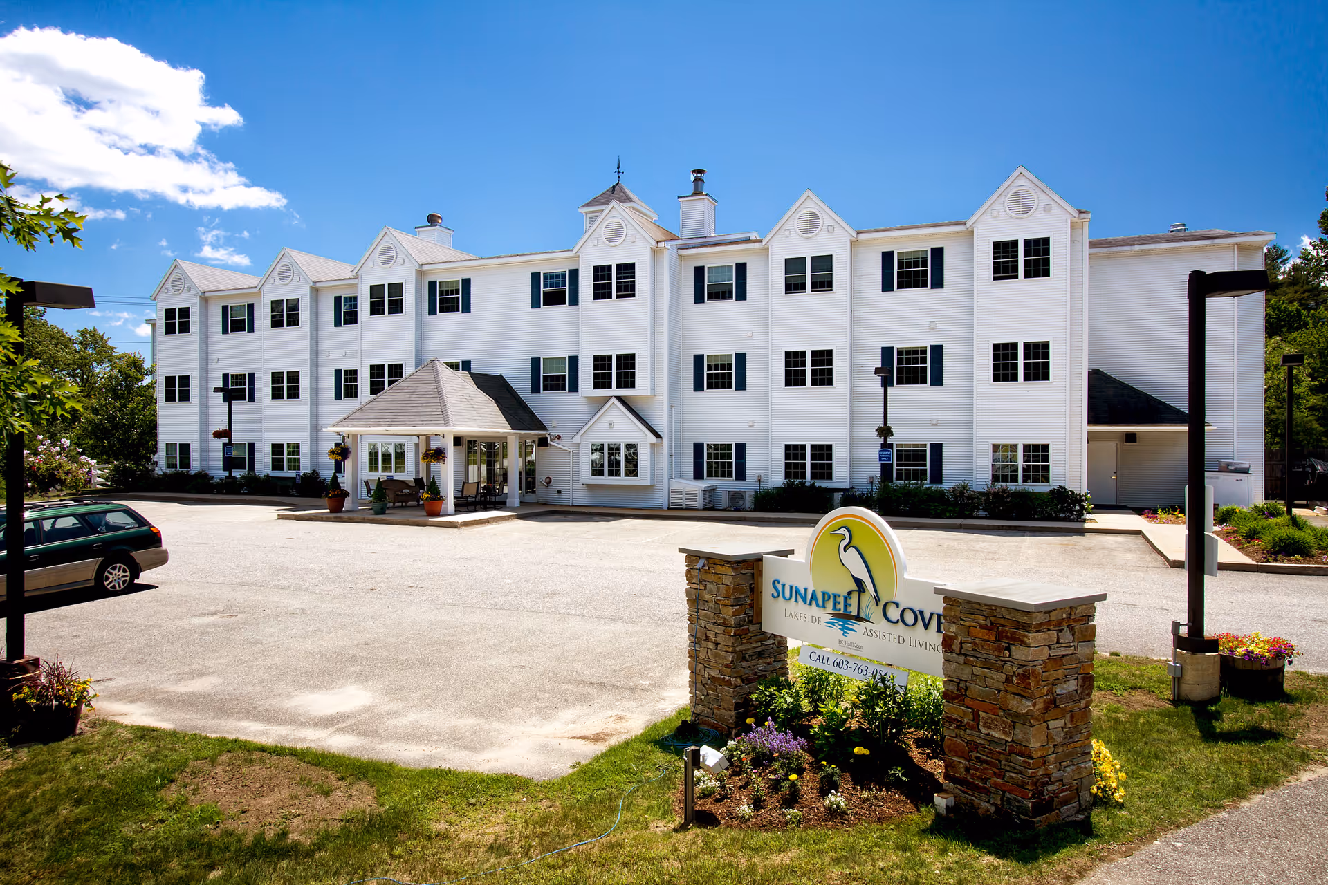 Exterior view of Sunapee Cove Assisted Living & Memory Care facility, a large white three-story building with multiple windows and a covered entrance. There is a parking area in front with a few cars and a landscaped sign with the facility's name and logo.
