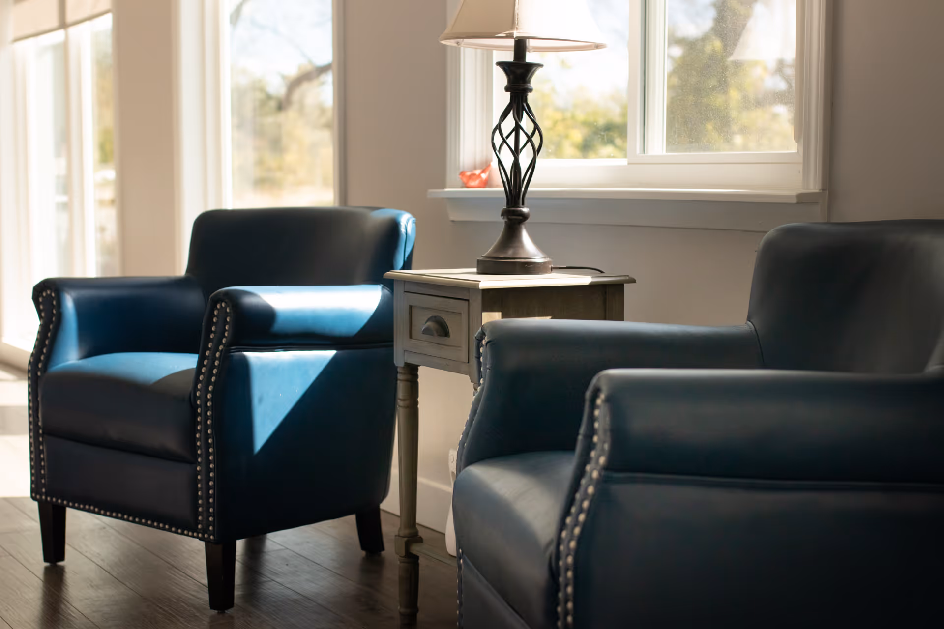 Two blue leather armchairs with nailhead trim positioned around a small wooden side table with a decorative lamp, next to a window letting in natural light.