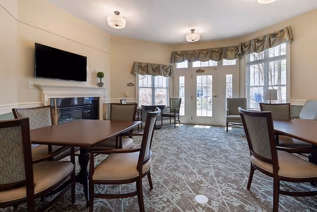 A well-lit common area with multiple wooden tables and cushioned chairs arranged on a patterned carpet. The room features large windows with valances, a glass door leading outside, a wall-mounted flat-screen TV above a white fireplace mantel, and neutral-colored walls.