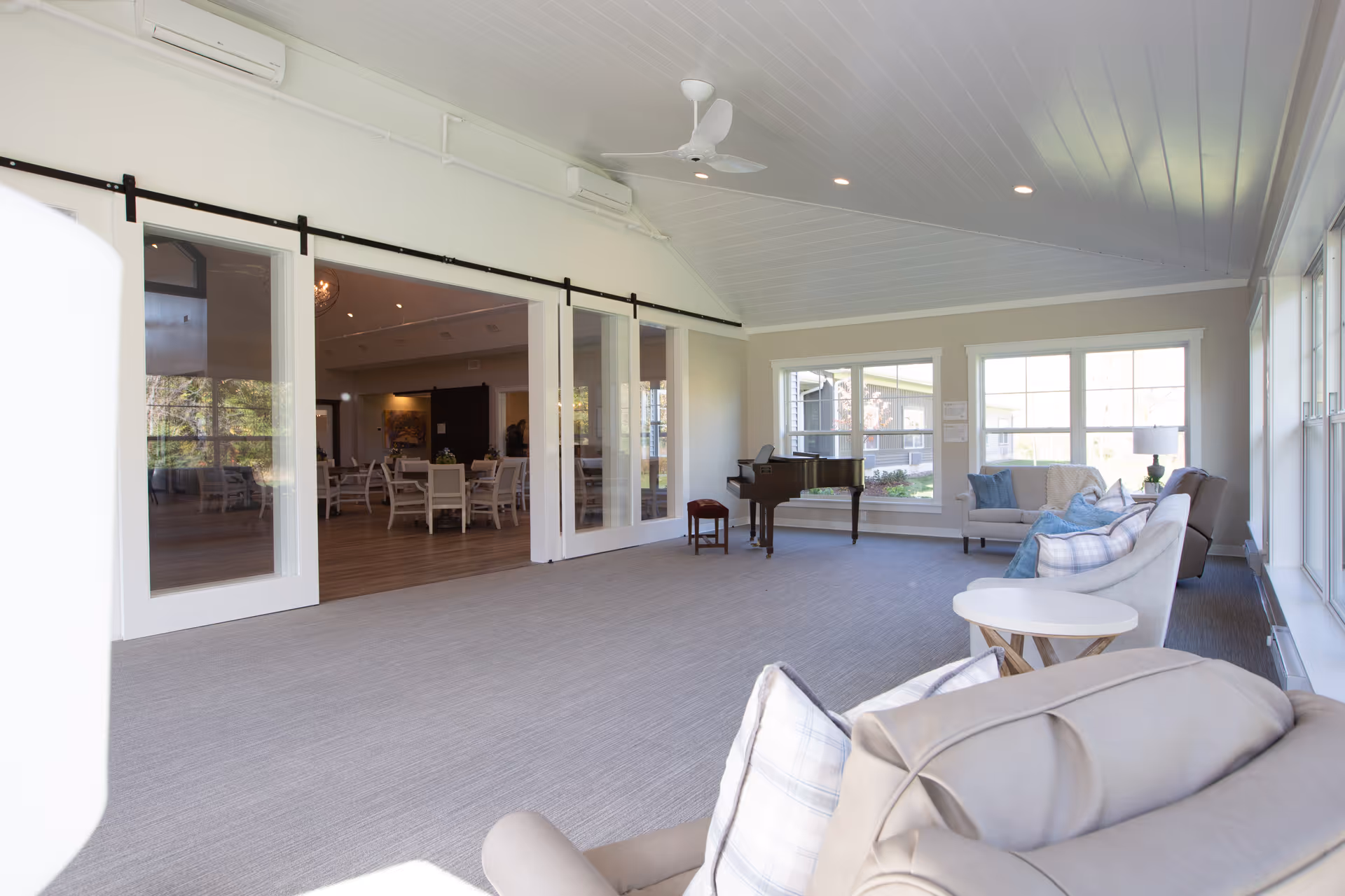 A bright and spacious living room area with large windows letting in natural light. The room features comfortable seating including armchairs and sofas with cushions, a small round side table, and a grand piano with a stool near the windows. Sliding glass doors lead to an adjacent dining area with tables and chairs.