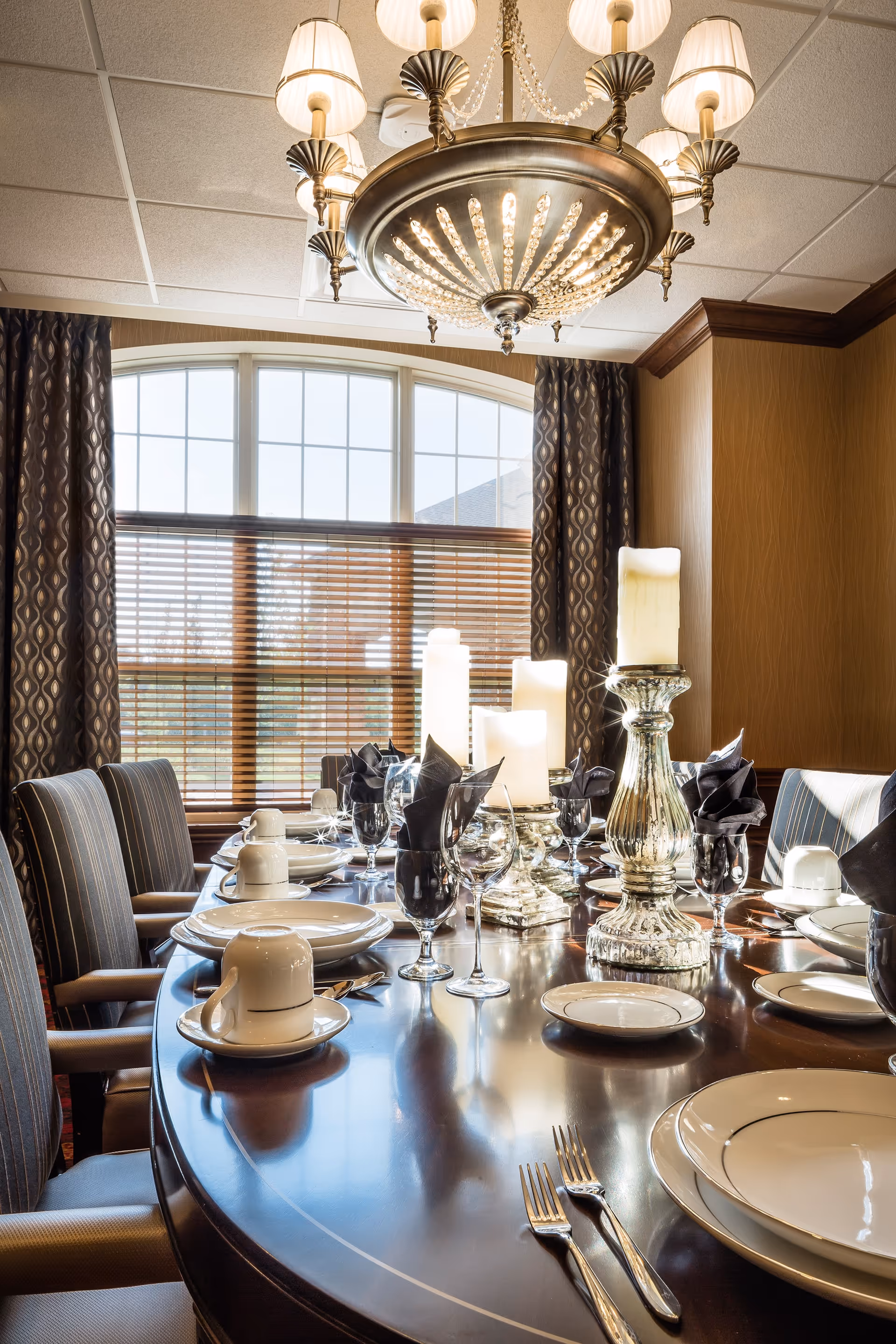 A formal dining room with a large wooden table set with white plates, cups, silverware, wine glasses, and black cloth napkins. The table is decorated with tall white candles in ornate silver candle holders. The room has large windows with wooden blinds and patterned curtains, and a decorative chandelier hangs from the ceiling.
