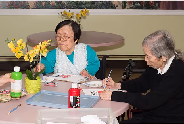 Two elderly women sitting at a round table engaged in painting activities with brushes and paper. One woman is wearing glasses and a turquoise sweater with a white vest, while the other woman is dressed in black. There are bottles of paint and a yellow orchid plant on the table. The background shows a beige wall and a floral painting.