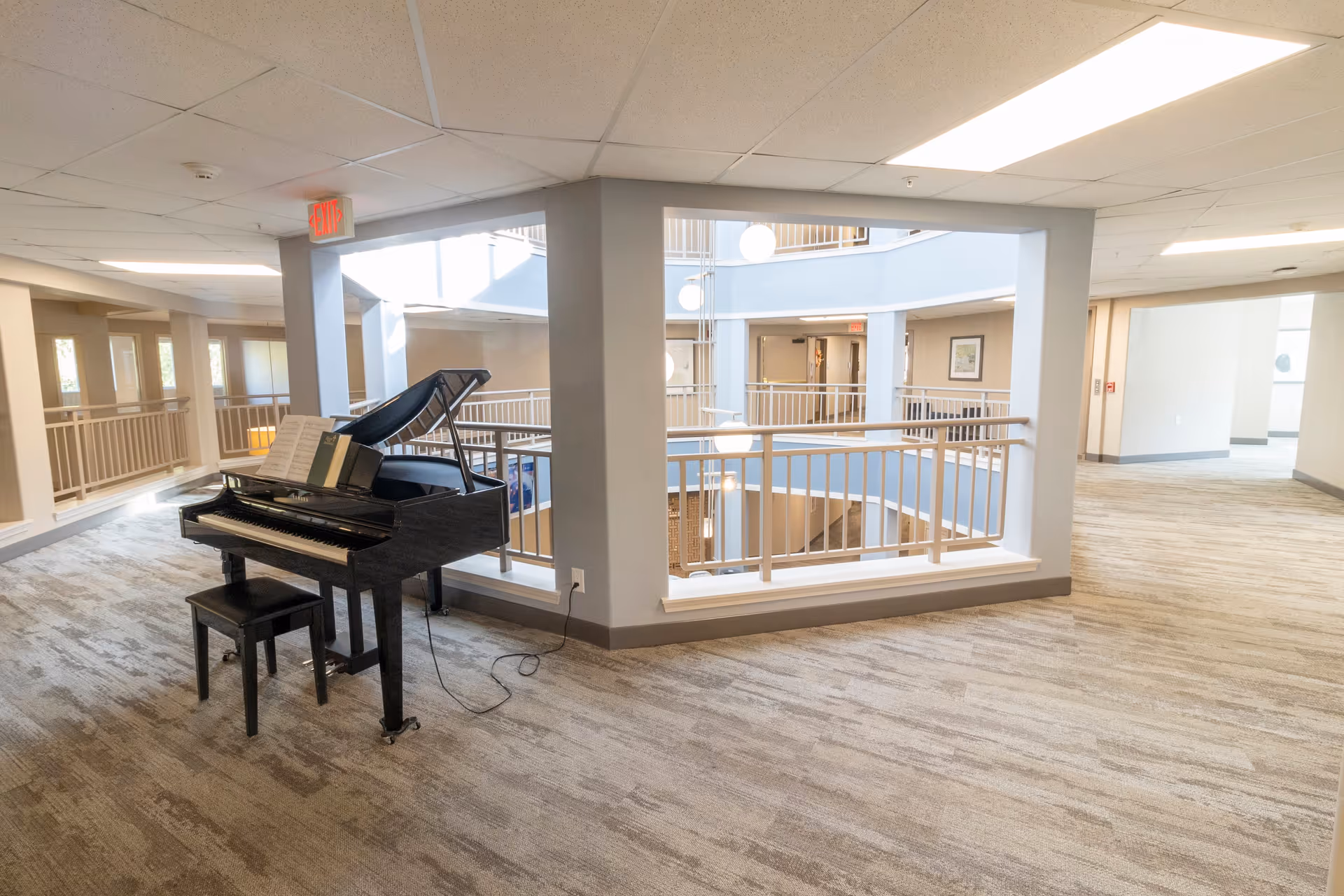 Interior view of a senior living facility hallway with a black grand piano and bench placed near a railing overlooking a lower floor. The area is well-lit with natural light coming through windows and ceiling lights. The carpet is patterned in neutral tones, and the walls are painted light gray.