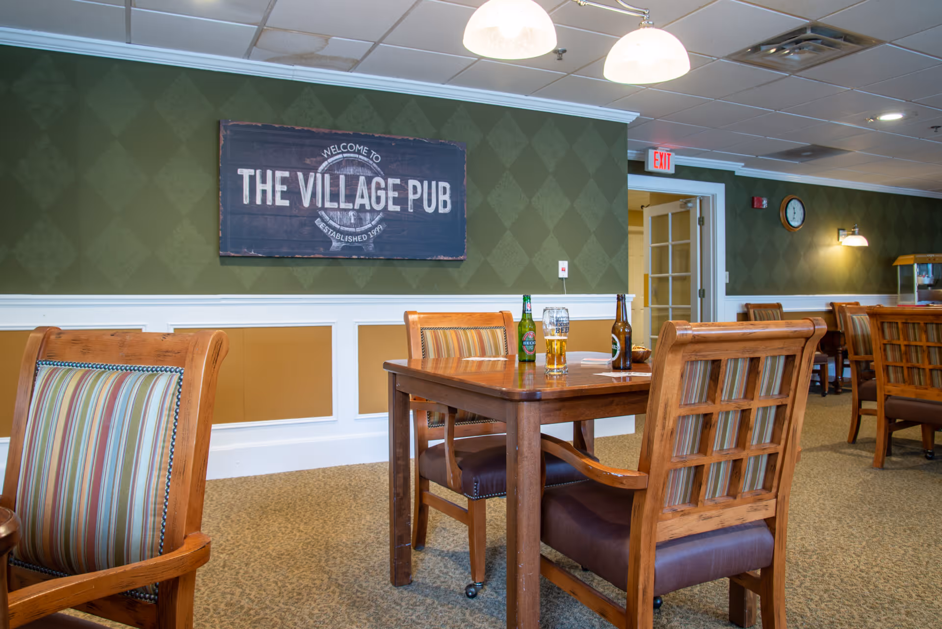 Interior view of The Village Pub at The Village at South Farms, featuring wooden tables and chairs with striped cushions, a green patterned wall with a sign that reads 'Welcome to The Village Pub Established 1987', and bottles and a glass of beer on the table.