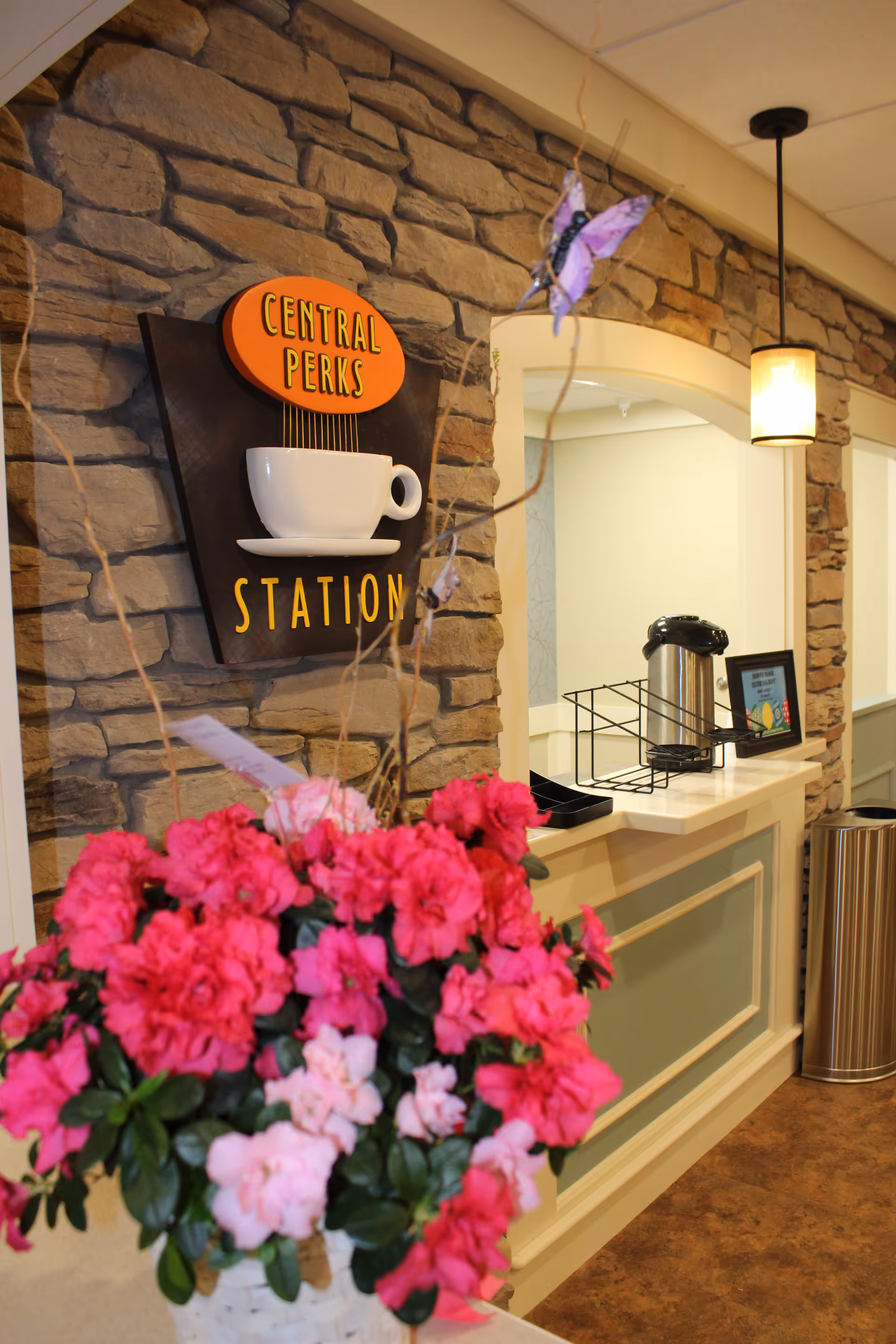 Interior view of a coffee station area named Central Perks Station with a stone wall backdrop, a hanging light fixture, a coffee dispenser on the counter, and a basket of vibrant pink and light pink flowers in the foreground.