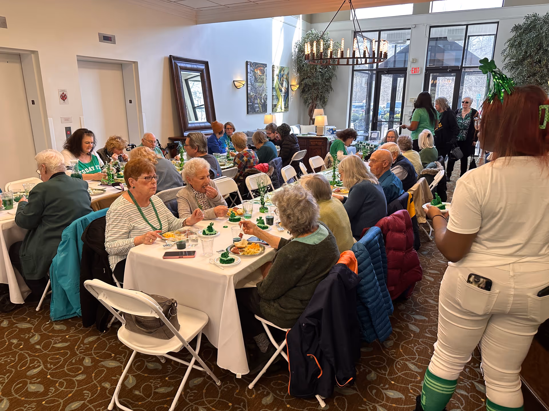 A group of older adults seated at long tables in a bright dining room enjoying a communal meal while staff and visitors stand nearby.