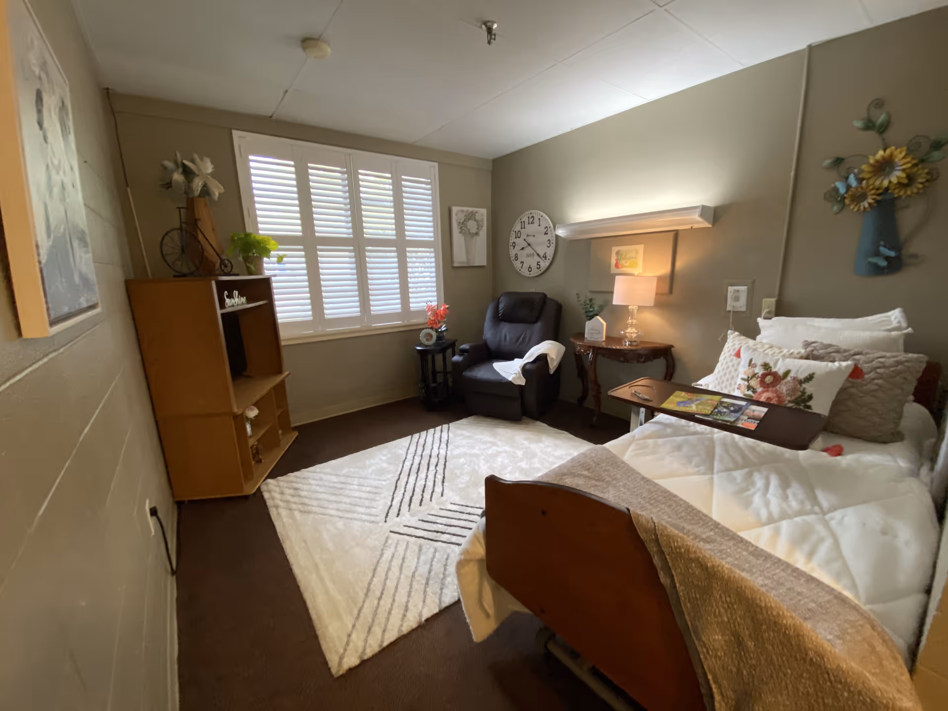 Cozy bedroom with a bed, recliner, side table, shelving unit and a window with shutters.