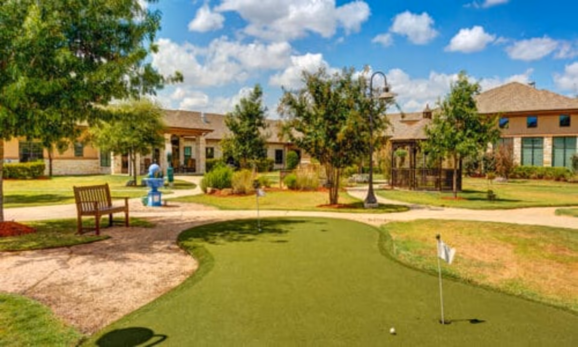 Outdoor view of a senior living facility with a small putting green, a wooden bench, trees, a blue water fountain, walking paths, and buildings in the background under a partly cloudy sky.