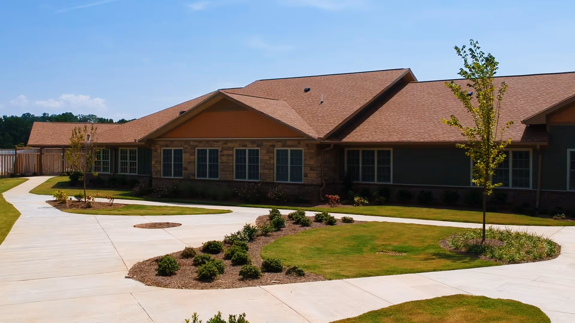 Single-story brick and siding senior living building with a circular concrete driveway and landscaped lawn under a blue sky.