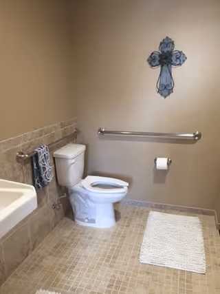 A clean bathroom with beige tiled floor and walls. There is a white toilet with a grab bar on the wall behind it and a toilet paper holder with a roll of toilet paper. A white sink is partially visible on the left side with a towel hanging on a grab bar. A decorative metal cross is mounted on the wall above the grab bar. A white bath mat is placed on the floor in front of the toilet.