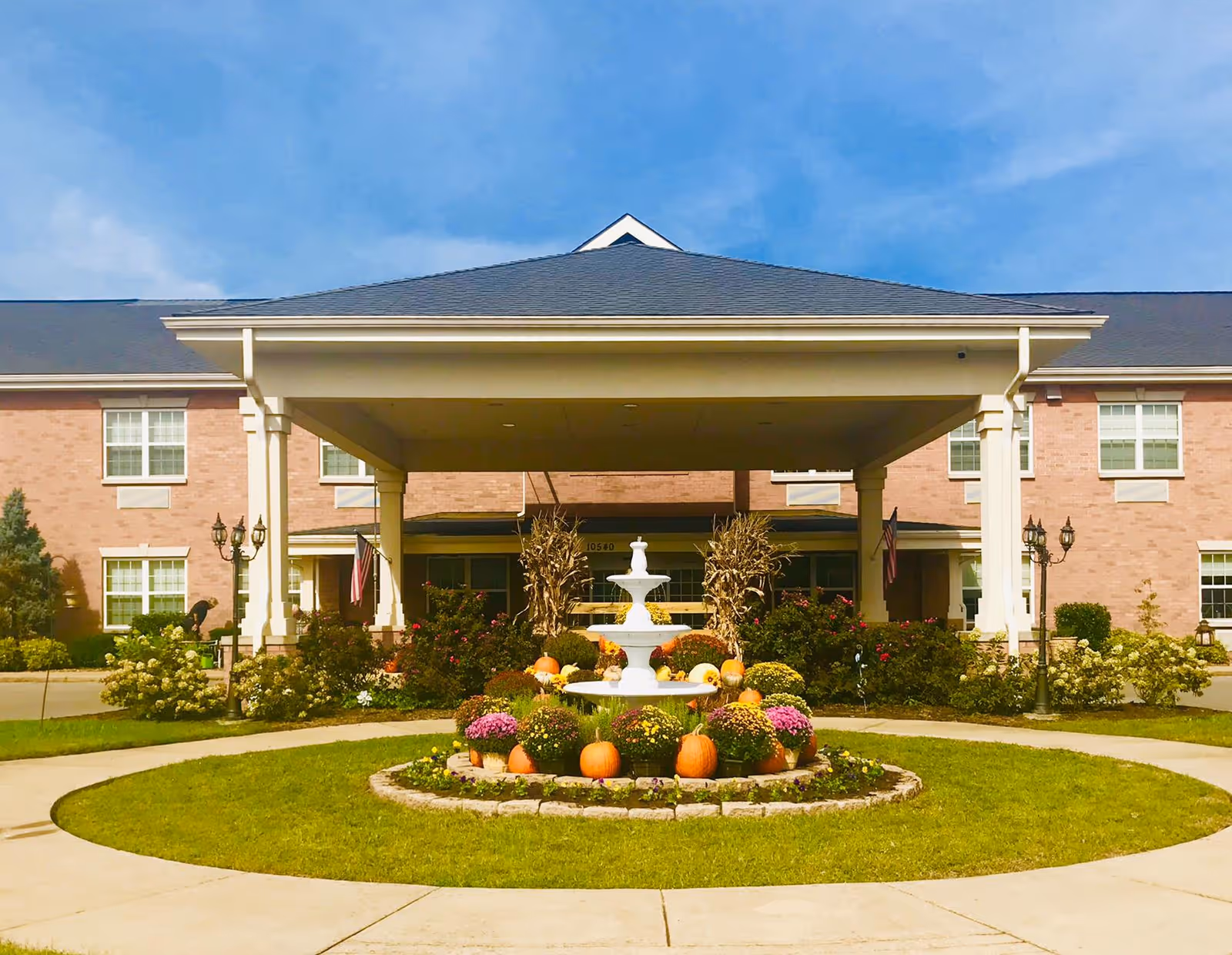 Front exterior view of The Pavilion Senior Living Smyrna building with a covered entrance, a circular driveway, and a landscaped roundabout featuring a white fountain surrounded by pumpkins and colorful flowers under a clear blue sky.