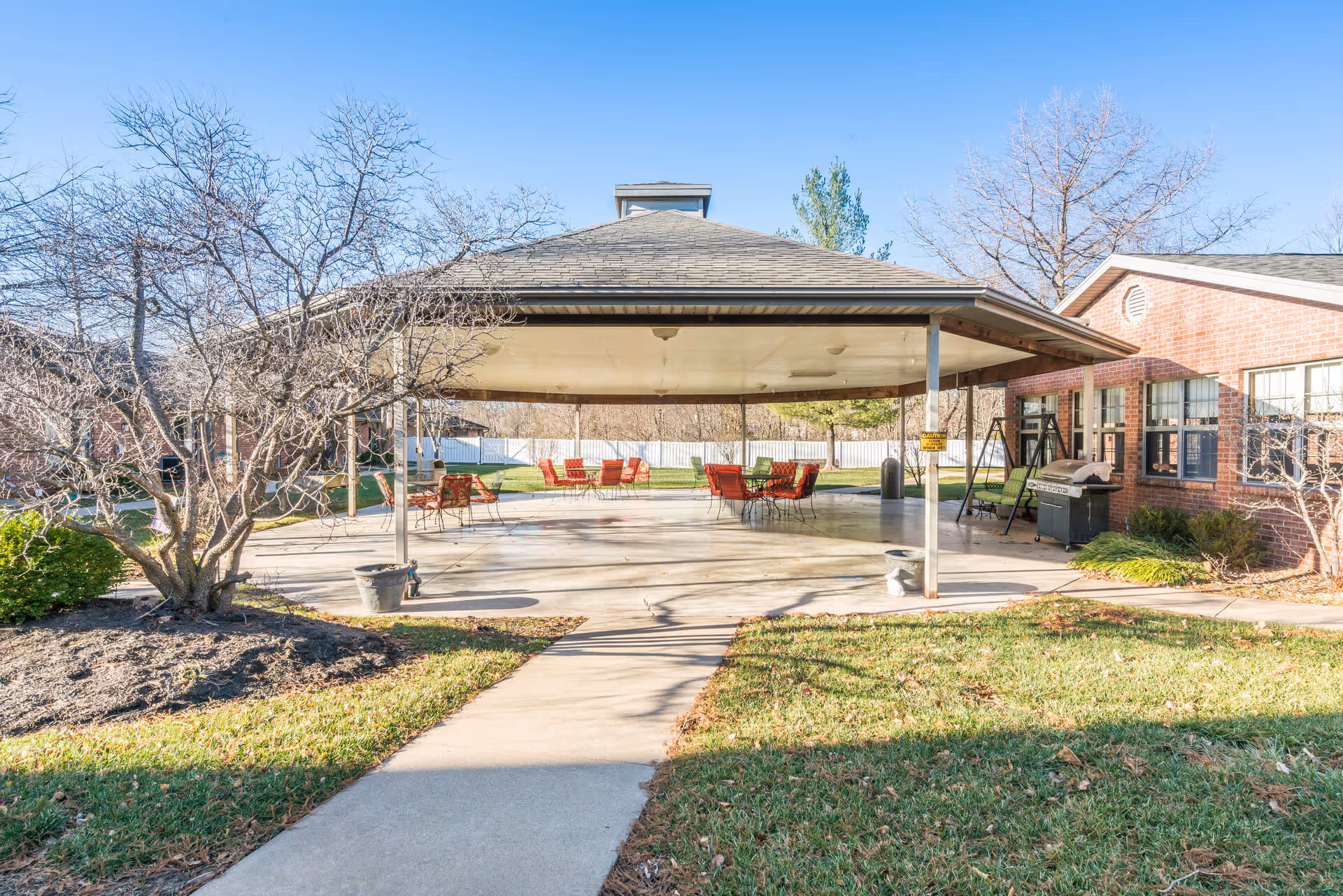 Outdoor covered patio area with red chairs and tables, adjacent to a brick building. There are leafless trees and green grass surrounding the paved walkway leading to the patio. A grill and swing bench are visible near the building.