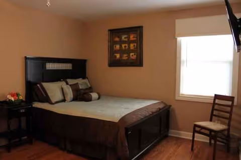 A furnished bedroom in Briar Hill Assisted Living with a queen bed, nightstand, window, and chair.