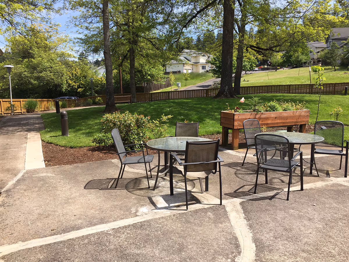 Outdoor patio area with two round glass tables and several metal chairs on a concrete surface. There is a raised wooden planter box with plants and flowers, surrounded by green grass, trees, and a wooden fence. Residential houses and a road are visible in the background under a clear sky.
