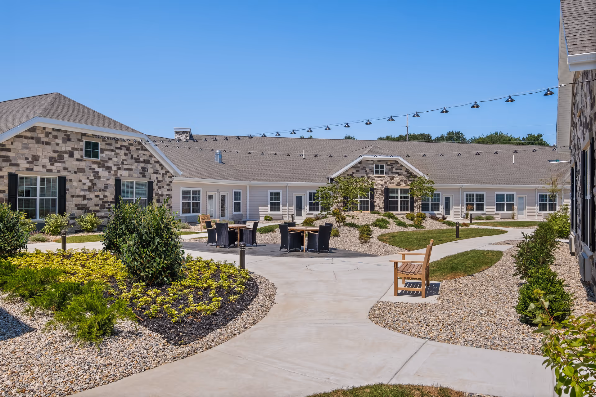 Outdoor courtyard area of a senior living facility with paved walkways, landscaped garden beds, shrubs, and small trees. There are several black wicker chairs and wooden tables arranged in the center, along with a wooden bench on the right side. The buildings surrounding the courtyard have a mix of stone and siding exteriors with multiple windows and doors. String lights are hung above the courtyard under a clear blue sky.