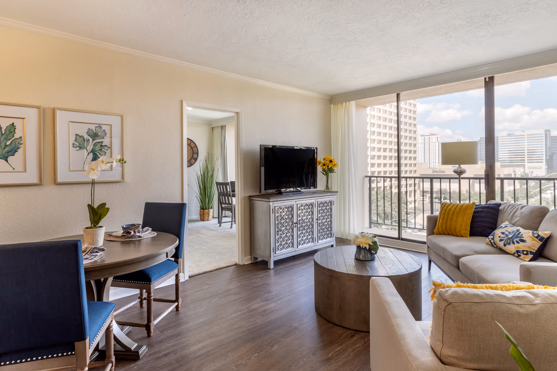 A bright living room with a large window showing a city view. The room features a beige sofa with colorful pillows, a round wooden coffee table with a flower vase, a TV on a decorative cabinet, and a small dining table with two blue chairs. There are two framed botanical prints on the wall and a doorway leading to another room with a large clock and a tall plant.