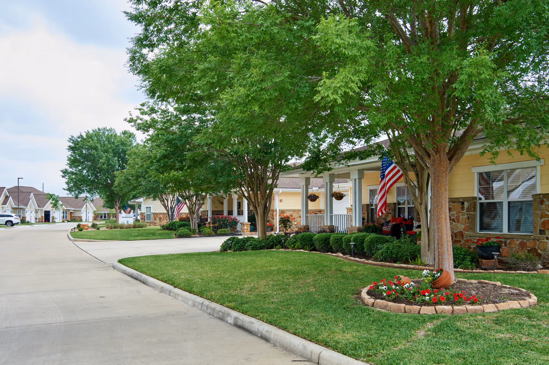 A peaceful residential street in The Village at Gleannloch Farms with well-maintained lawns, trees, and flower beds. Single-story houses with stone and yellow siding, front porches decorated with American flags and hanging flower baskets line the street. The sky is partly cloudy.