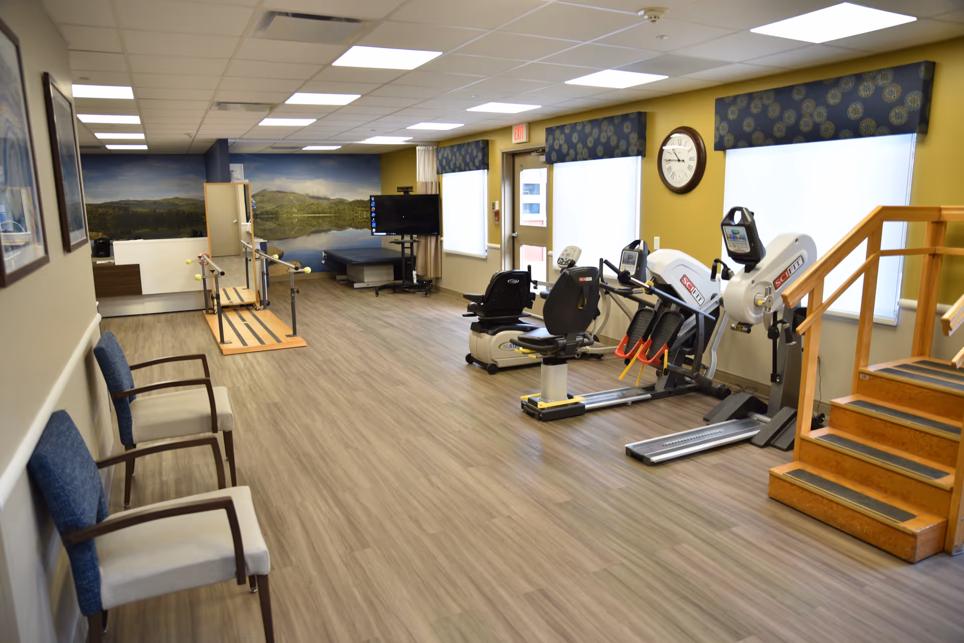 Bright rehabilitation and exercise room with cardio machines, parallel bars, practice stairs, chairs, and a wall clock.