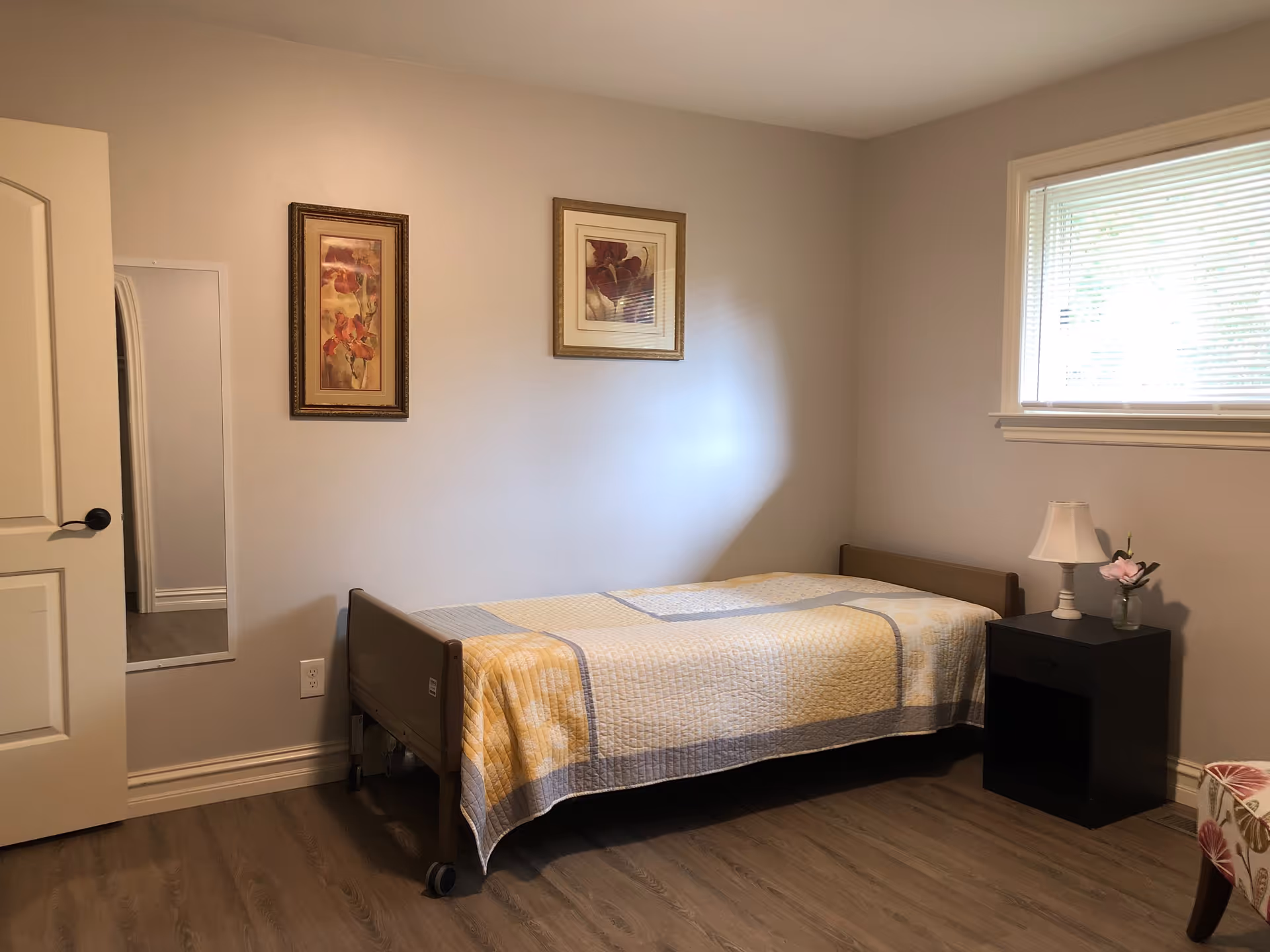 A simple bedroom with a single bed covered with a yellow and gray quilt. There are two framed floral pictures on the wall above the bed. A small black nightstand next to the bed holds a white lamp and a small vase with a pink flower. A window with blinds is on the right wall, and a door is partially open on the left side. The floor is wood, and there is a full-length mirror mounted on the wall near the door.